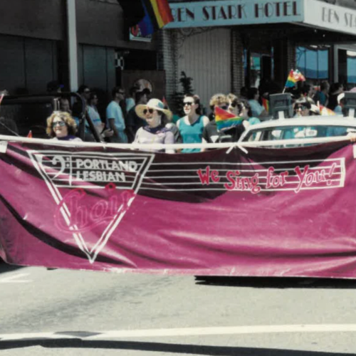 Group of people holding a Portland Lesbian Pride banner with rainbow flags in an outdoor setting