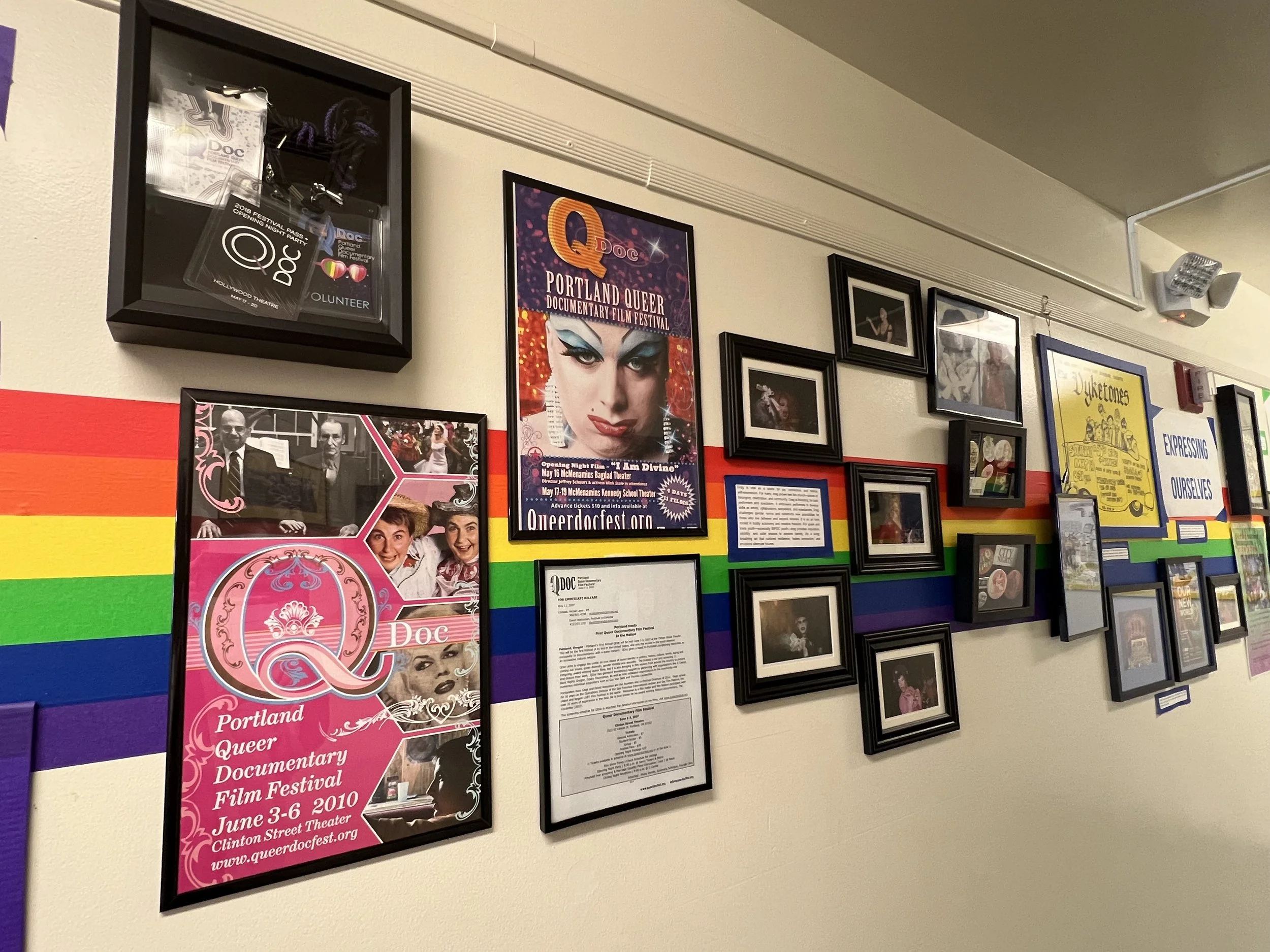 Wall decorated with photographs, posters, and framed documents, including a poster for the Portland Queer Documentary Film Festival.