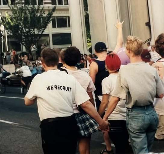 A group of young people holding hands and raising their hands during a public event or parade on a city street, with large columns and buildings in the background.