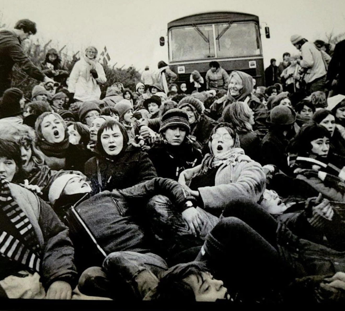 Black and white photo of a large crowd of young people, many sitting or lying on the ground, some with mouths open, appearing to shout or sing, with a bus in the background.