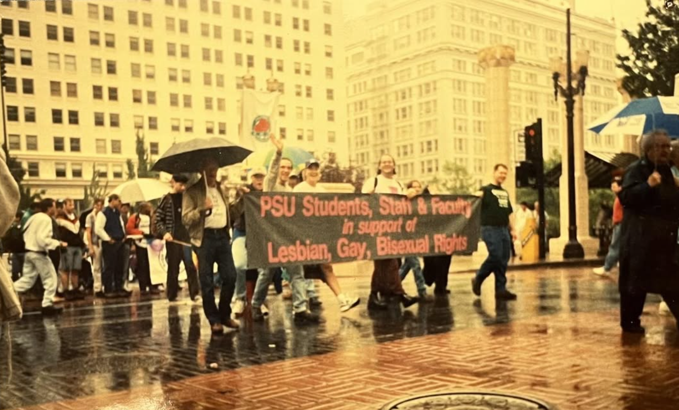 Group of people participating in a march or demonstration on a rainy day, holding a banner that reads 'PSU Students, Staff & Faculty in support of Lesbian, Gay, Bisexual Rights,' with some holding umbrellas and standing on wet pavement near tall buil