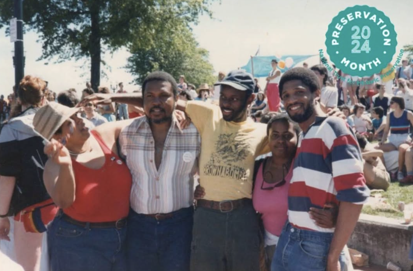 Group of five diverse people standing outdoors at a festival or gathering, with many people and trees in the background.