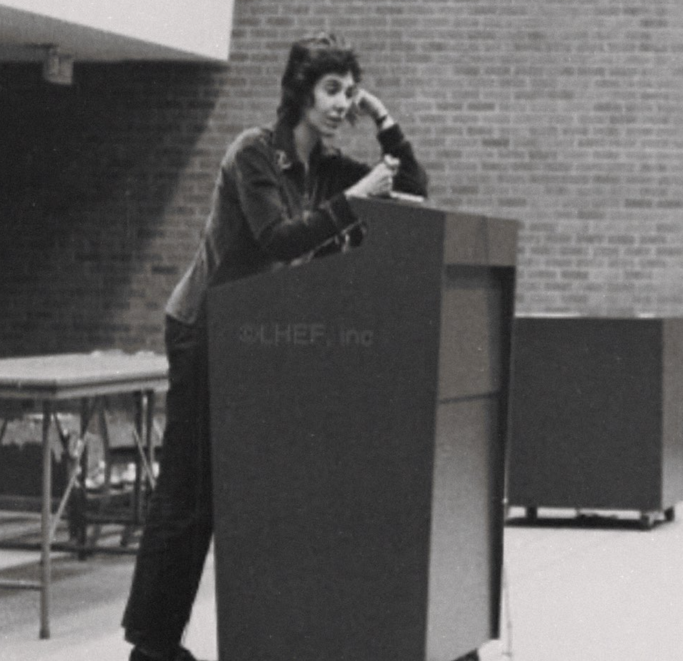 A woman standing behind a lectern with a microphone, speaking or giving a presentation, in a room with brick walls.