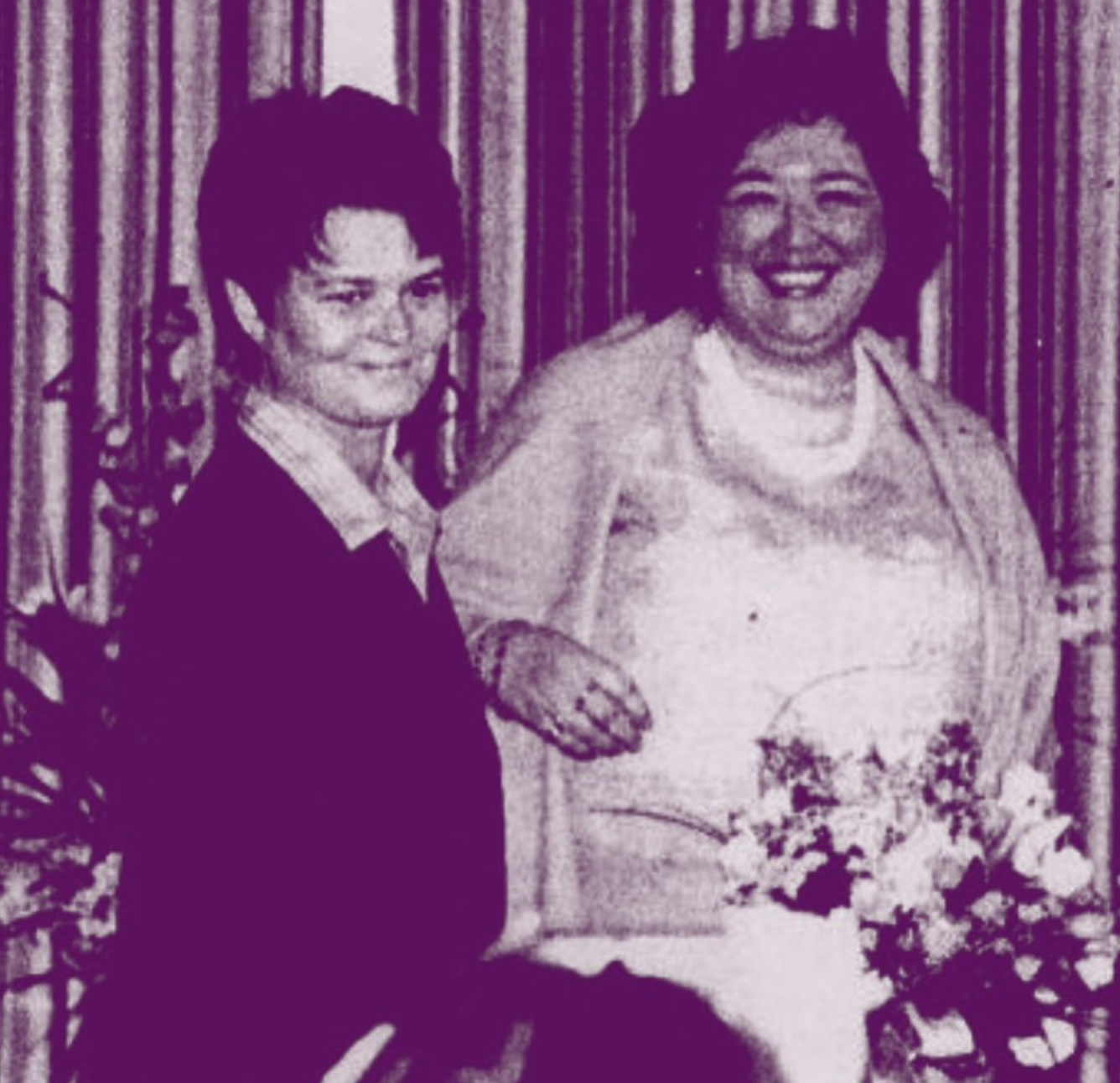 Two women standing close together, smiling. One woman is wearing a dark uniform and glasses, the other is dressed in a light-colored blouse and cardigan, holding a bouquet of flowers, with a backdrop of vertical curtains.
