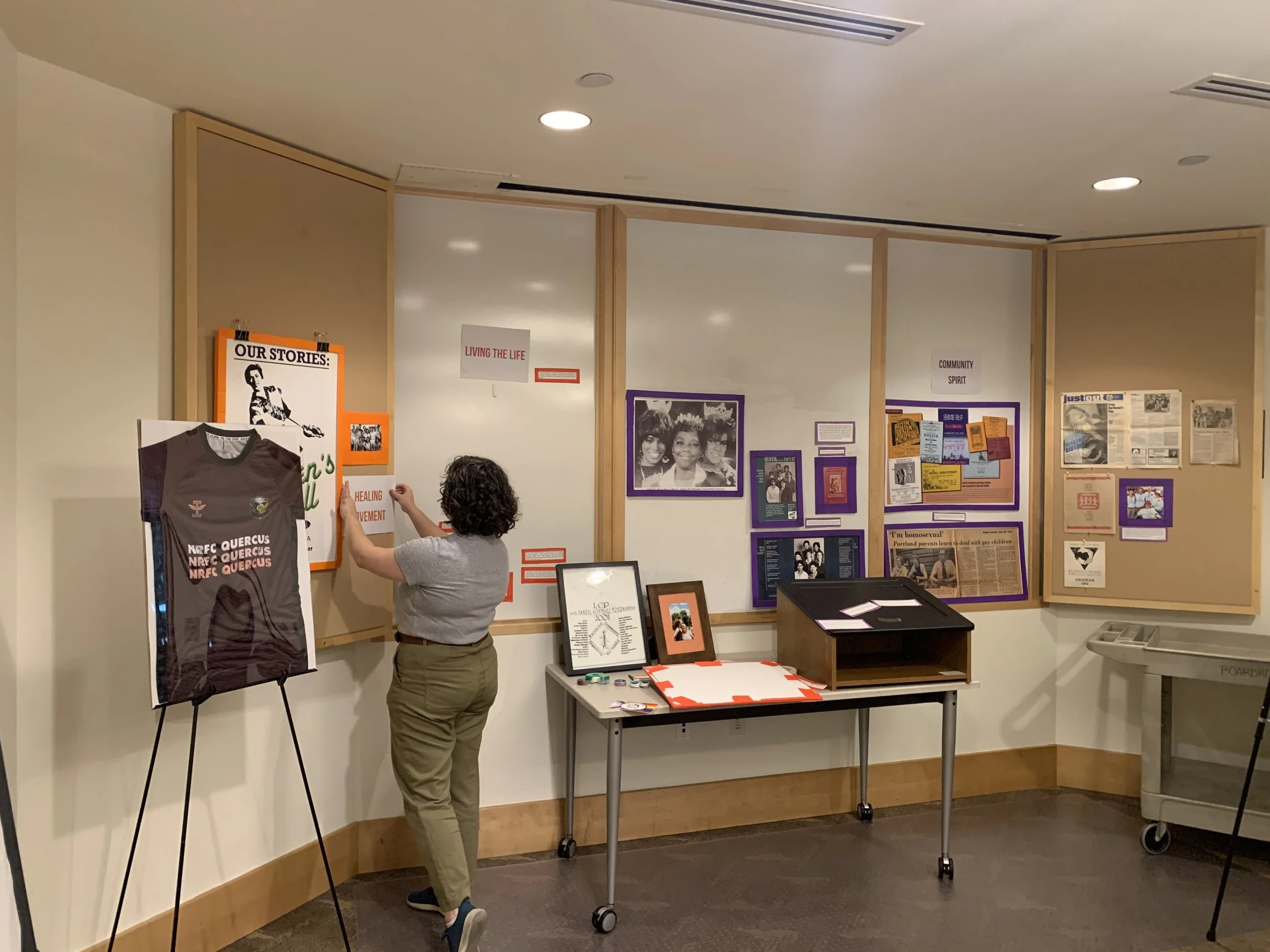 A person stands in front of an informational display on a wall, with posters, framed photographs, and written materials relating to community, history, or social issues, in an indoor setting with art and informational displays.