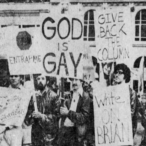 Protesters holding signs with messages such as "God is Gay," "Give Back the Column," and "Write O. Brian" in front of a brick building.