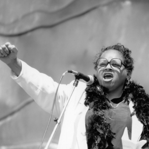 Woman with glasses and curly hair speaking passionately into a microphone, wearing a white jacket and black feather boa, with her fist raised.