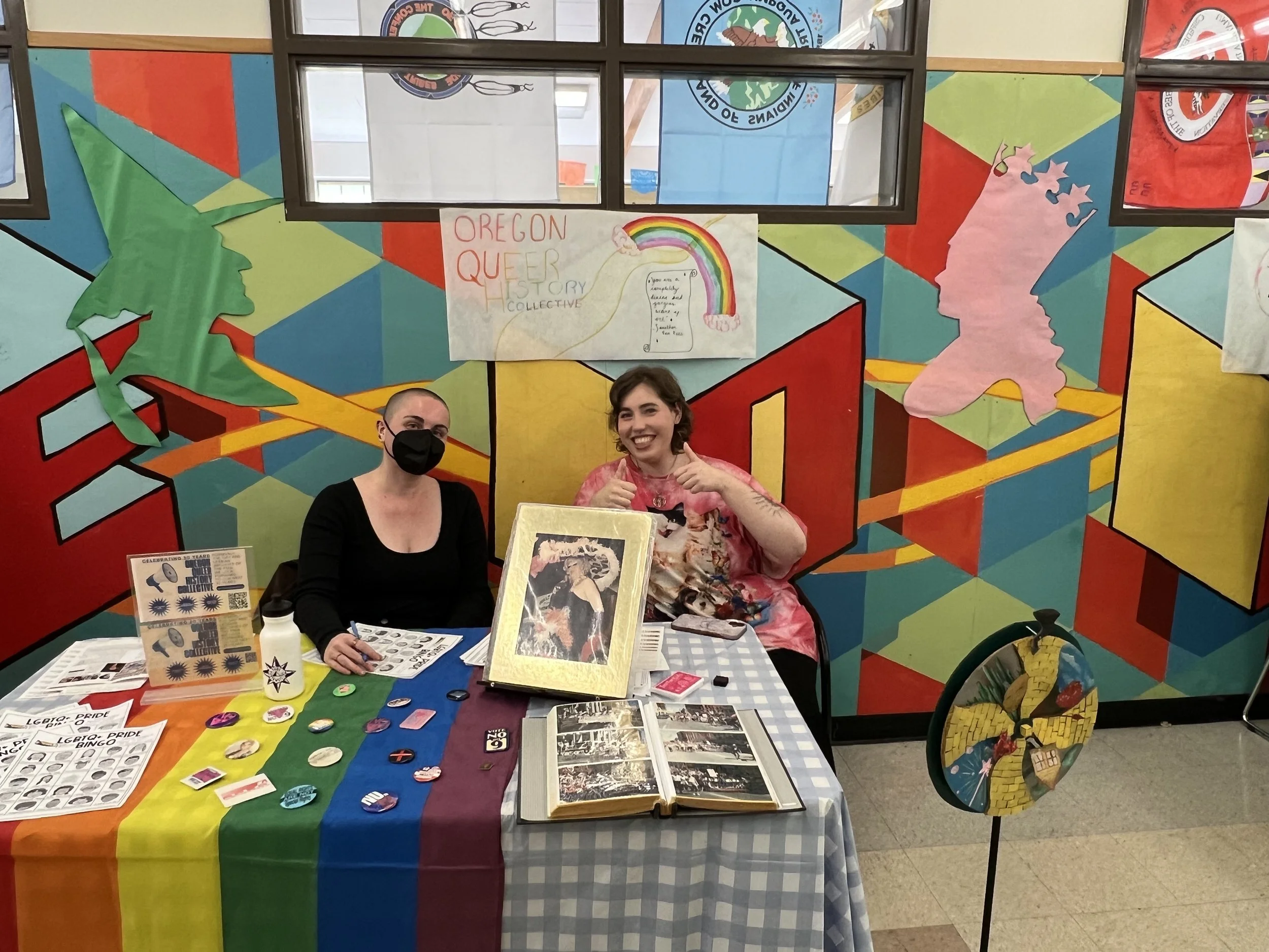 Two people sitting behind a table with LGBTQ+ Pride items, in front of a colorful mural featuring geometric shapes, a green tree, and a pink silhouette of a woman's face with star-shaped earrings, and a sign that reads 'Oregon Queer Story Collective' with a rainbow drawing.