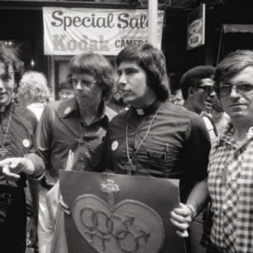 Group of young people at a protest or rally, holding a flag with interconnected gender symbols, in front of a sign that says 'Special Sale.'