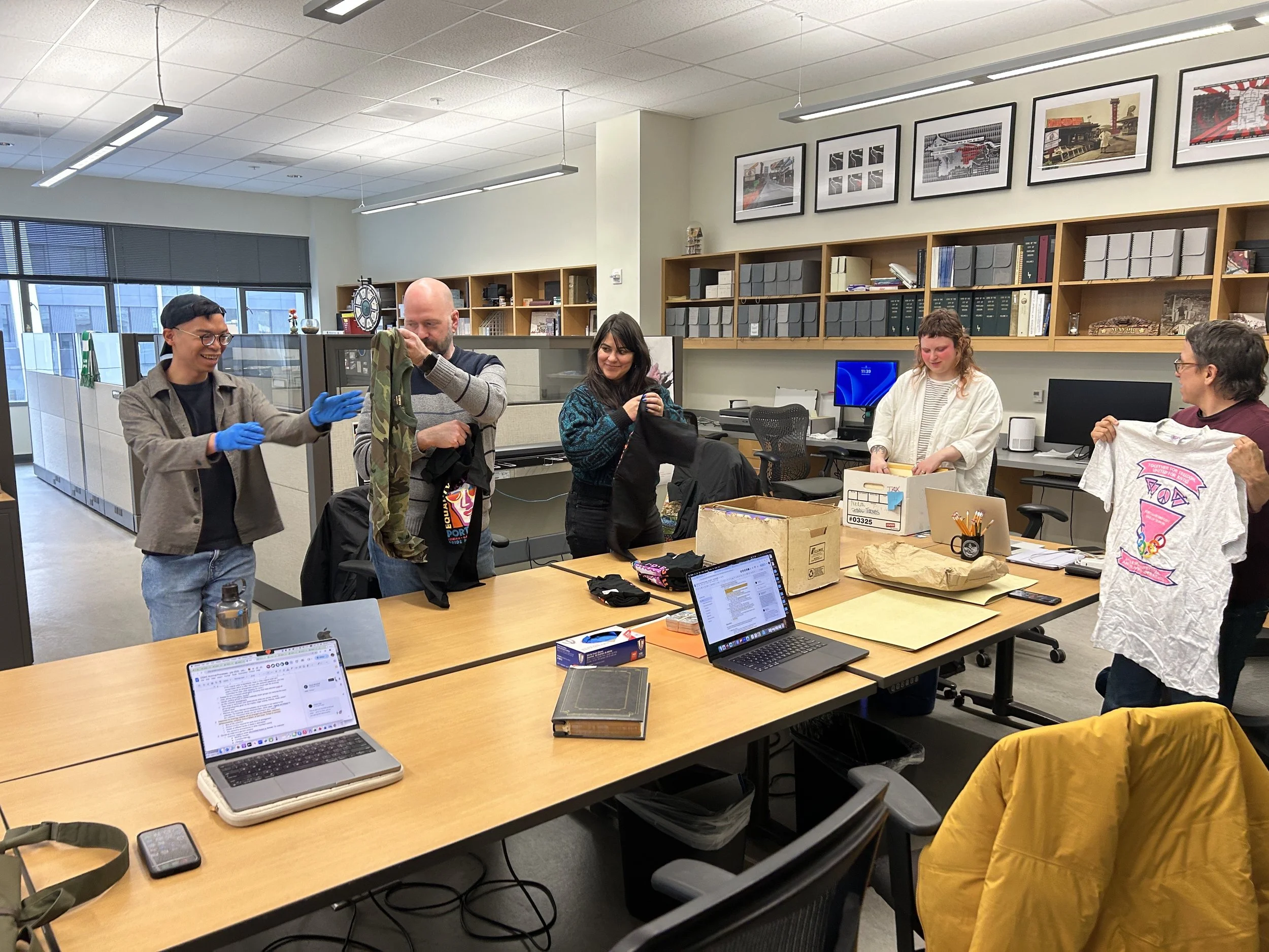 People gathered around a conference table in an office, some are smiling, and engaging in conversation, with laptops, papers, and boxes on the table.