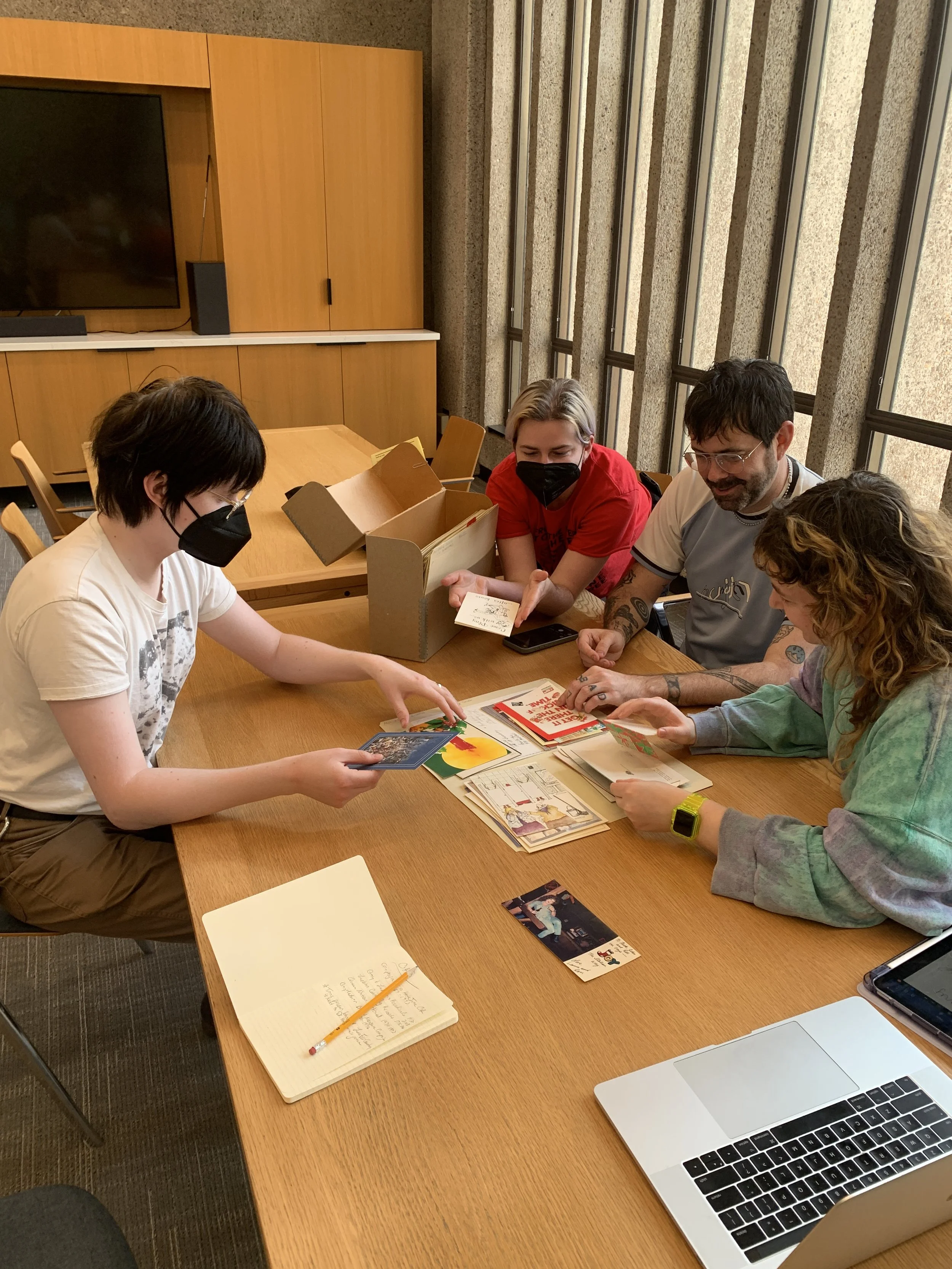 Group of four people sitting around a wooden table looking at cards, some using masks, with a box of cards in the center, a notebook, a pen, and an open laptop on the table.