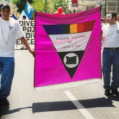 Two people holding a pink flag with a black, yellow, and red stripe at the top, and a black triangle with a white circle containing a black outline of the state of Oregon. The flag has Spanish text that reads 'Poder Latino de Oregon.'