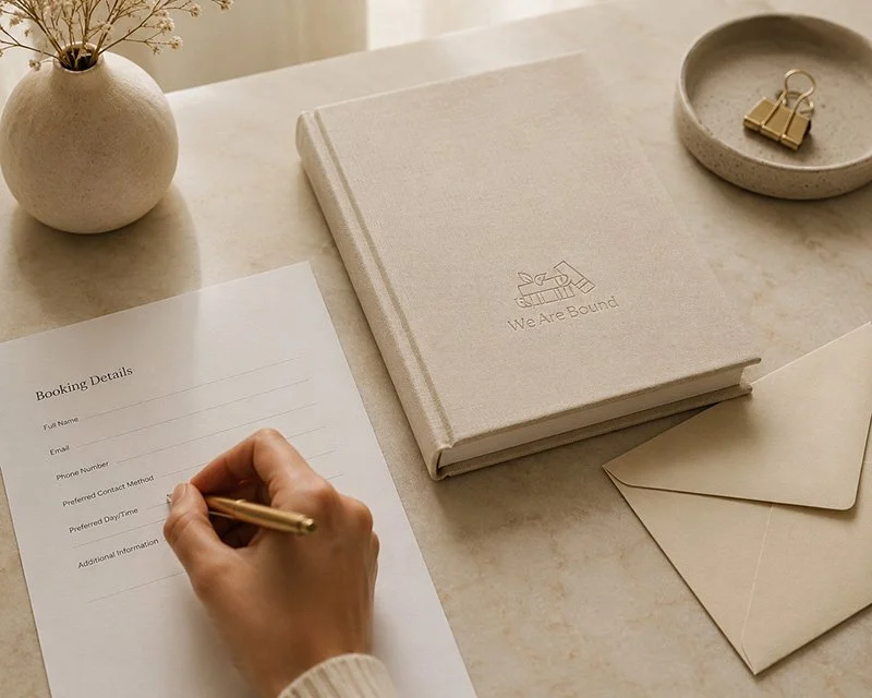 A person filling out a booking details form on a desk with a book titled 'We Are Bound,' an envelope, a small dish with gold binder clips, and a round vase with dried flowers.