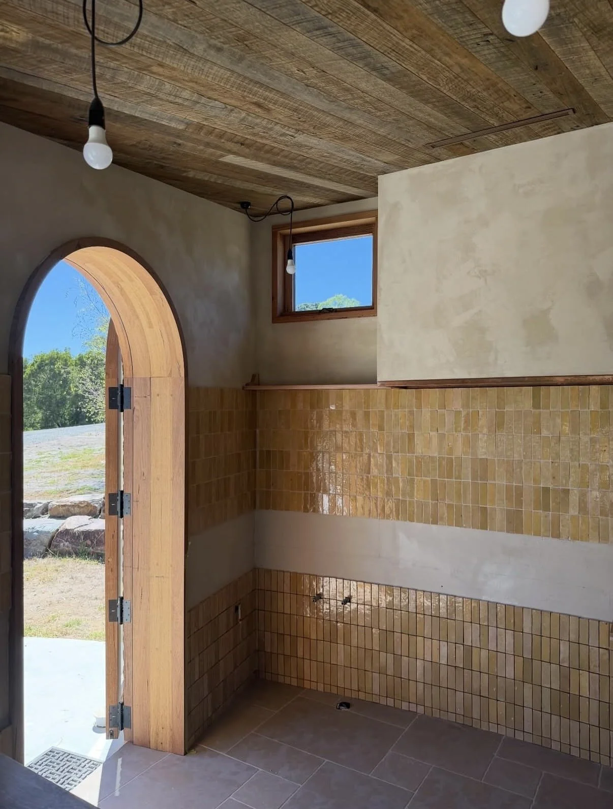 Interior of a room under construction with a wooden arched doorway, small window, tiled walls, and light fixtures hanging from the ceiling.
