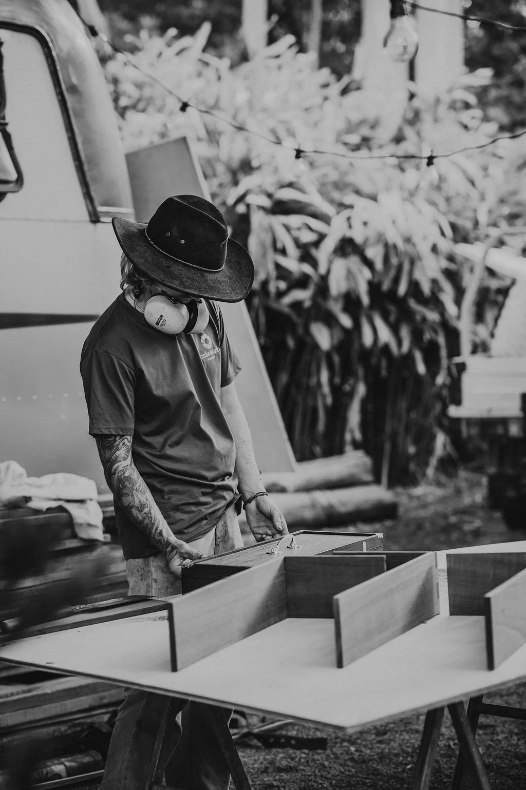 A person wearing a wide-brimmed hat and headphones, with tattoos on their arms, is working on a woodworking project outdoors near a camper trailer, with plants and trees in the background.