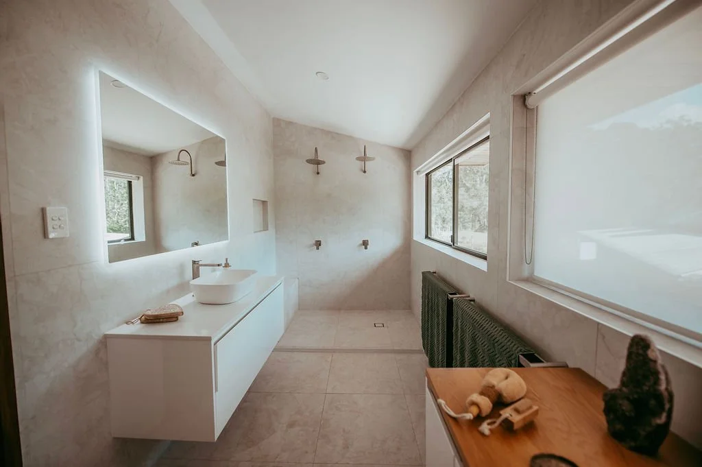 Modern bathroom with a large mirror, white vanity, vessel sink, two shower heads, large window, and natural light.