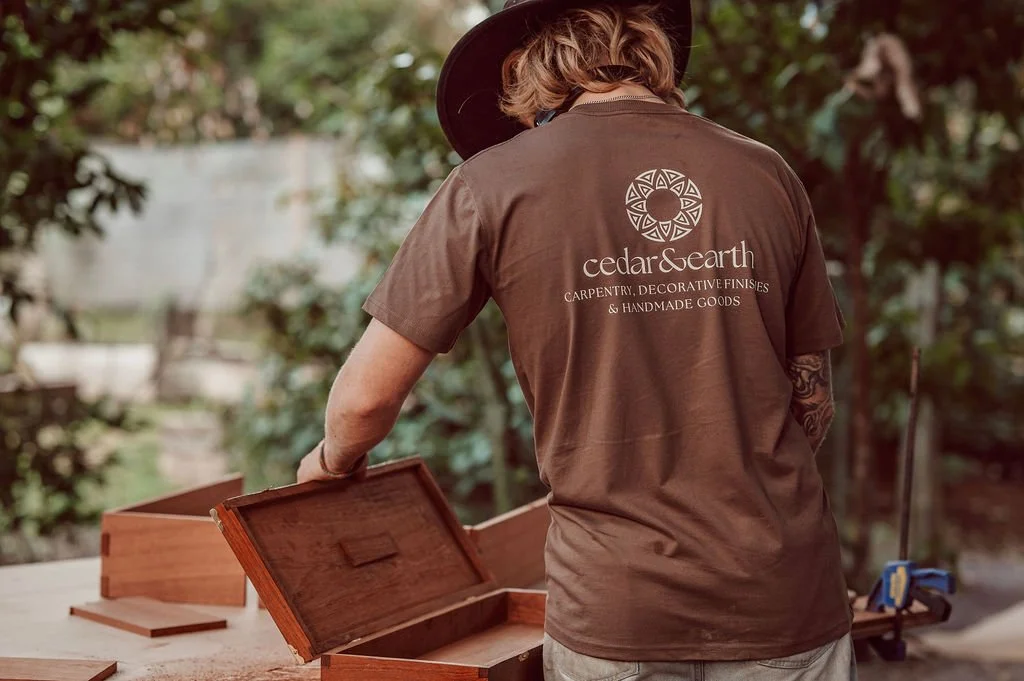 Person working on woodworking project outdoors, wearing a brown t-shirt with 'cedar & earth' logo, surrounded by woodworking tools and pieces of wood.