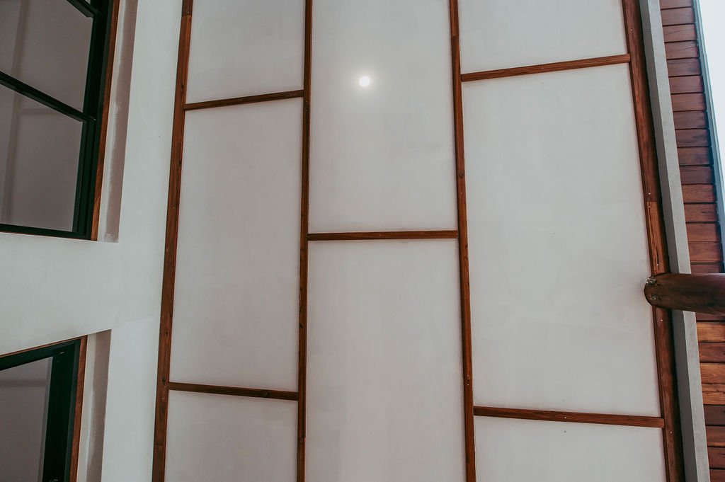 Ceiling with wooden beams and drywall panels, with part of a window frame visible on the left.