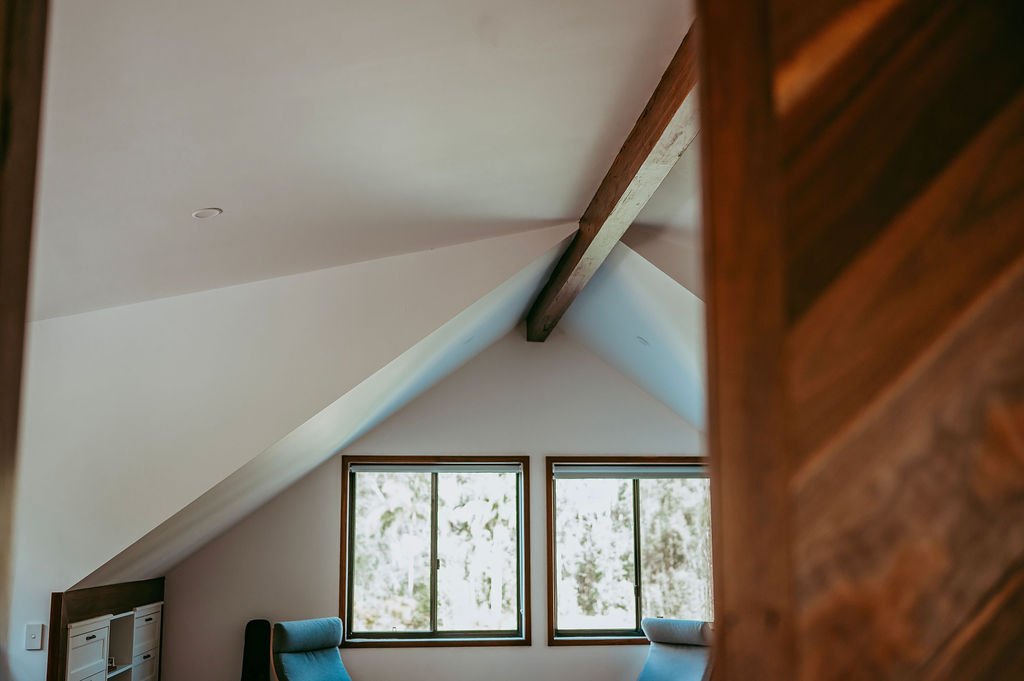 View of an attic room with sloped ceiling, two windows showing trees outside, and part of a wooden cabinet on the right.