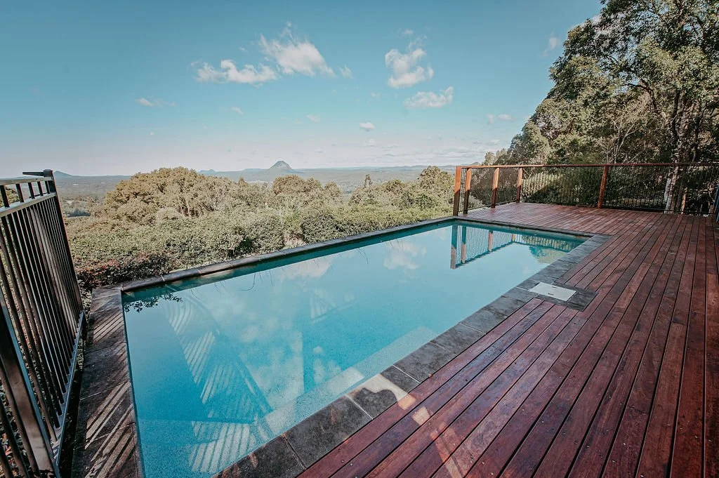 A small rectangular swimming pool with clear water on a wooden deck, overlooking a scenic landscape of trees and distant mountains under a partly cloudy sky.