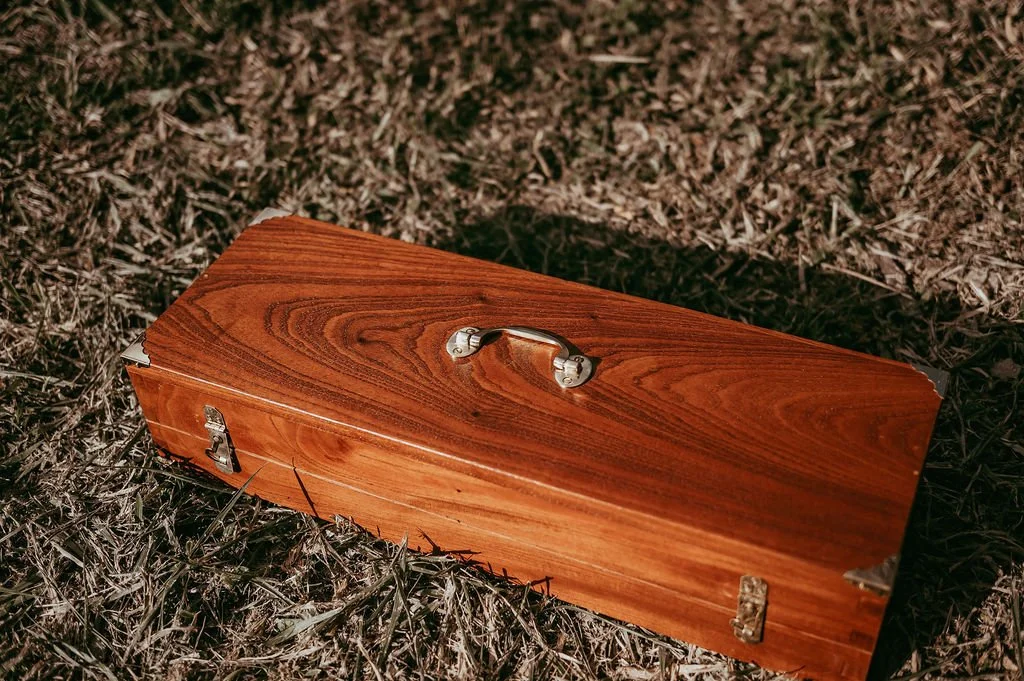 A wooden coffin with a metal handle lying on dry grass.