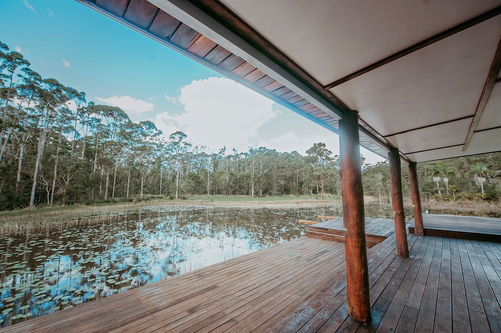 View of a wooden deck and covered patio overlooking a pond surrounded by trees with lily pads and a partly cloudy sky.