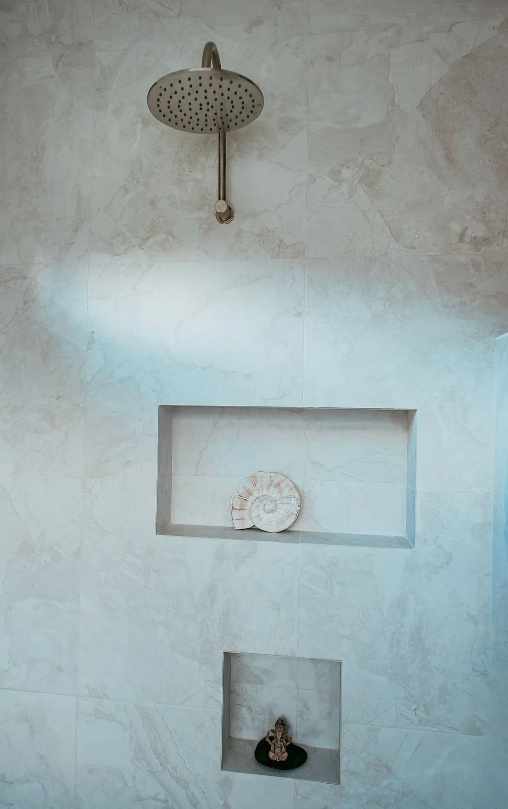 Shower with a round rainfall shower head, a built-in shelf with a decorative shell, and a smaller niche with a statue, all on a marble wall.