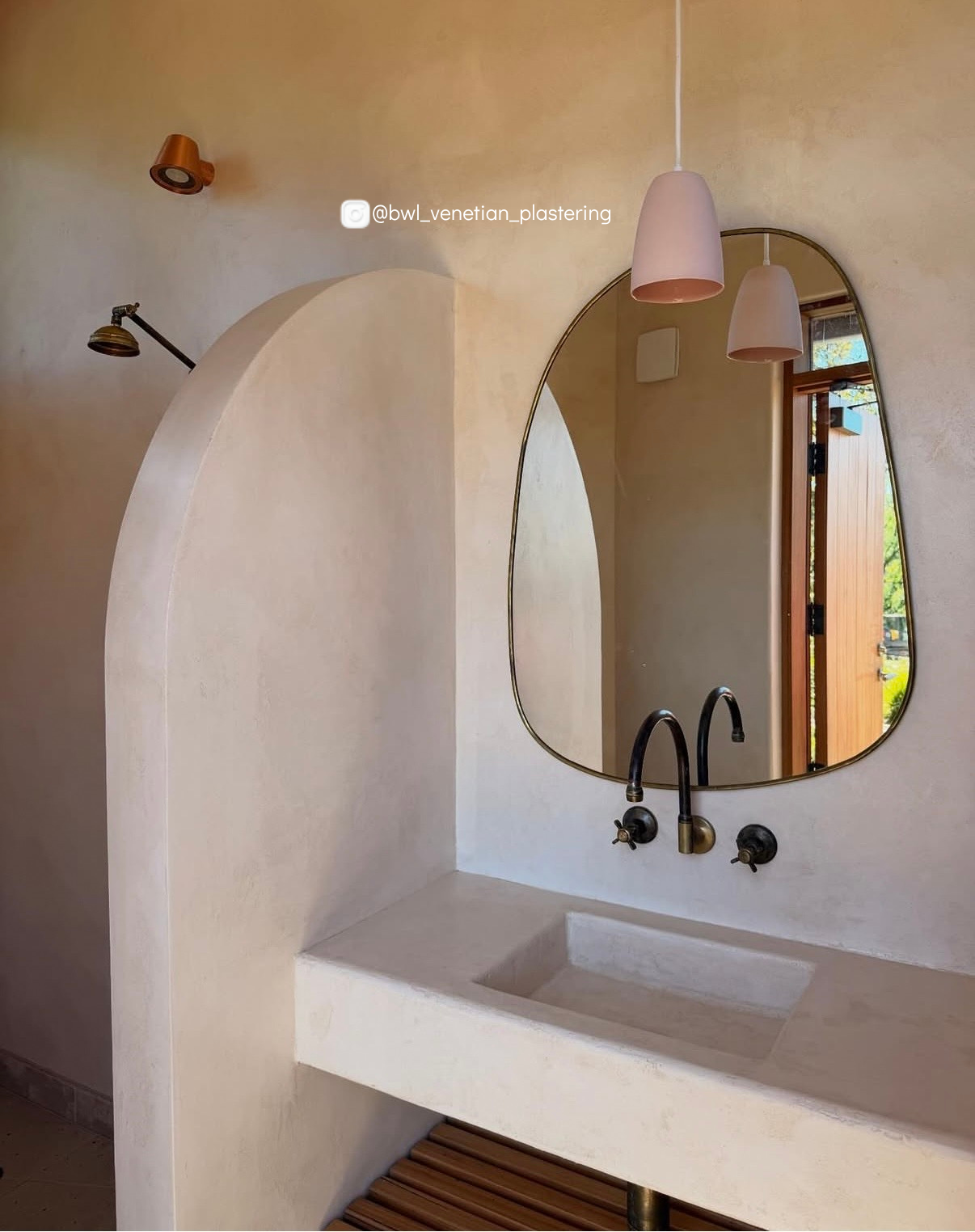 Minimalist bathroom with a curved arch wall, oval mirror, black faucet, white sink, pink pendant lights, and a wooden door reflected in the mirror.