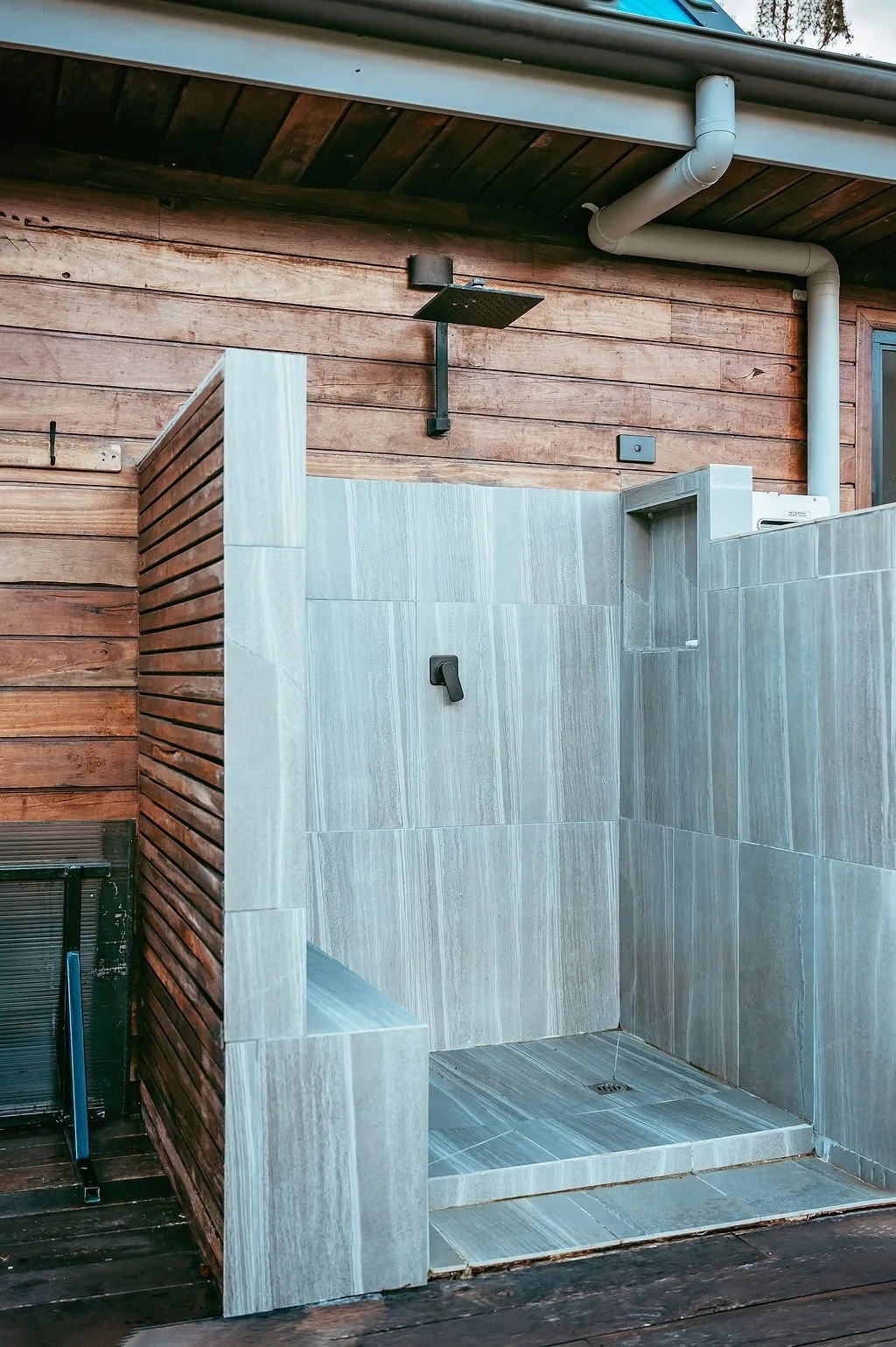 An outdoor shower area with a rain shower head mounted on a wooden wall, alongside a wall with a black handle, surrounded by a partial wooden screen, with visible roof and drain pipes above.