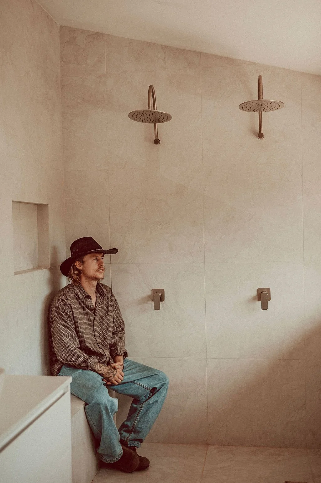 A man with tattoos, wearing a cowboy hat, plaid shirt, and jeans, sitting in a shower room under two empty showerheads on a beige tiled wall.