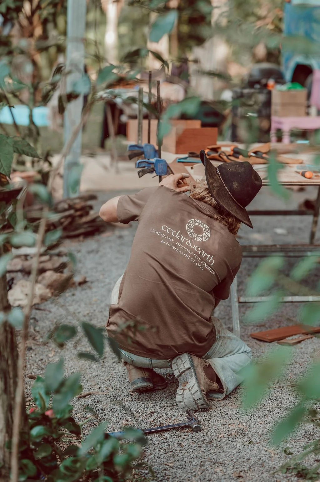 Person kneeling on gravel ground, wearing a hat and brown shirt, working with tools at an outdoor woodworking or carpentry project surrounded by trees and woodworking supplies.