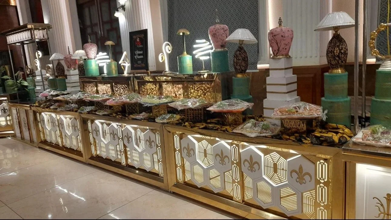 A buffet table with various wrapped food items, decorative lamps, and ornate containers, set in an elegant room with gold and white accents and patterned walls.