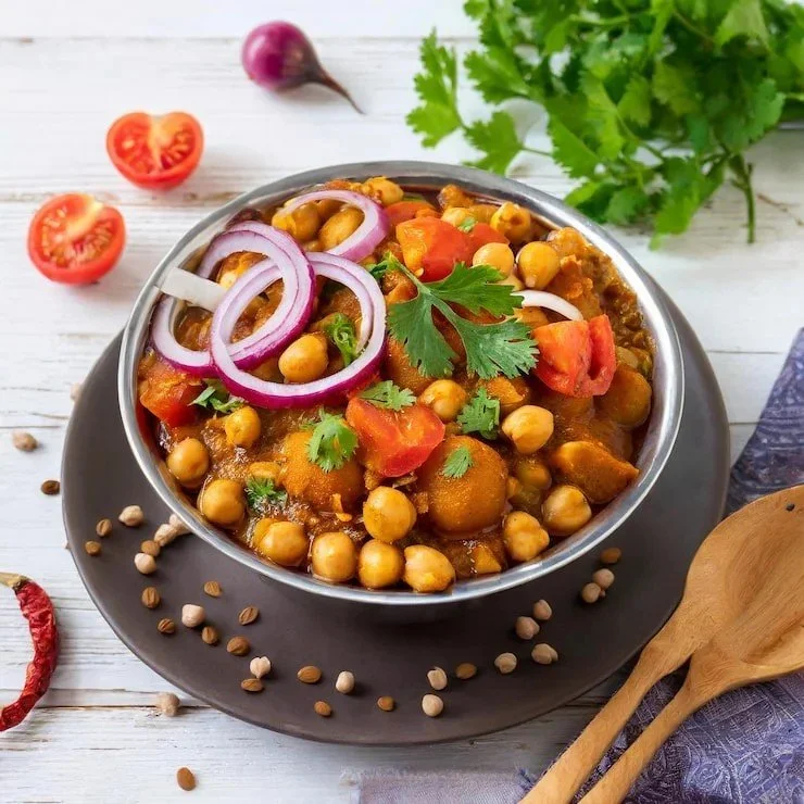 A bowl of chickpea curry garnished with sliced red onions and cilantro, served on a dark plate with scattered chickpeas and vegetables around it on a white wooden table.
