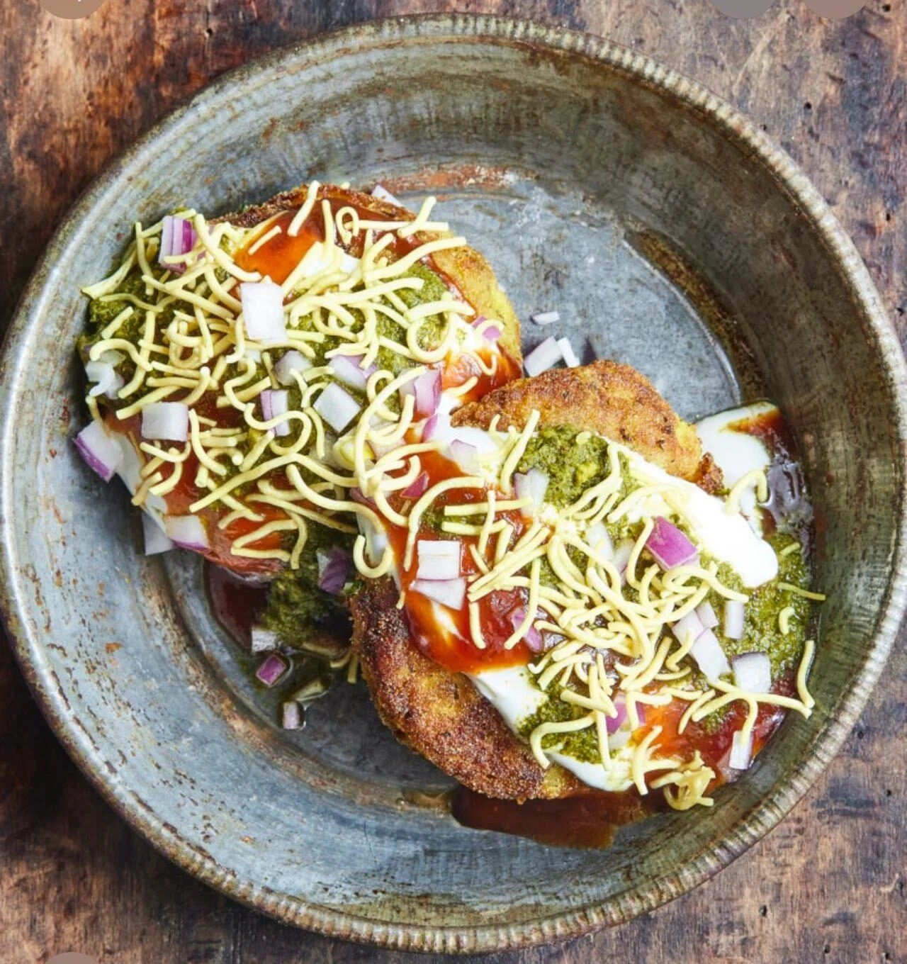 Close-up of a plate with two stuffed bread rolls topped with shredded cheese, chopped onions, green sauce, and hot sauce, on a rustic wooden surface.