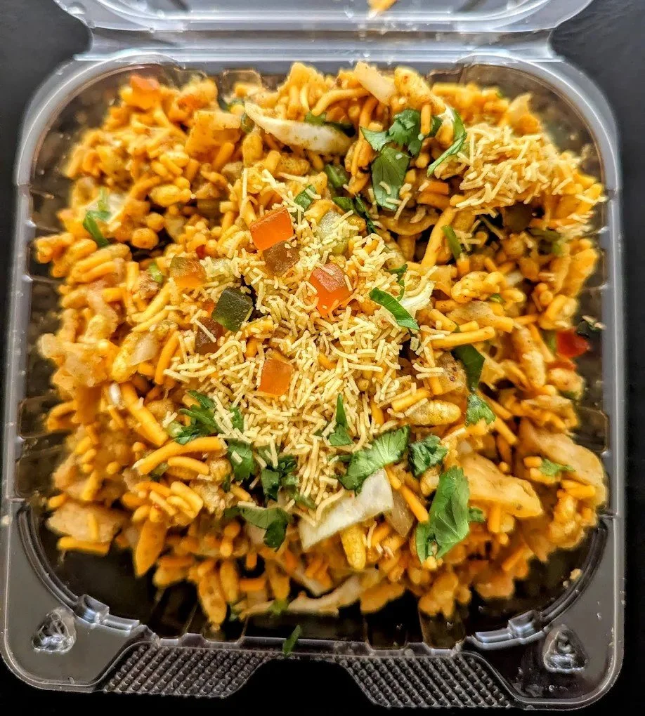 Takeout container of stir-fried noodles with vegetables, topped with shredded cheese and garnished with chopped cilantro.