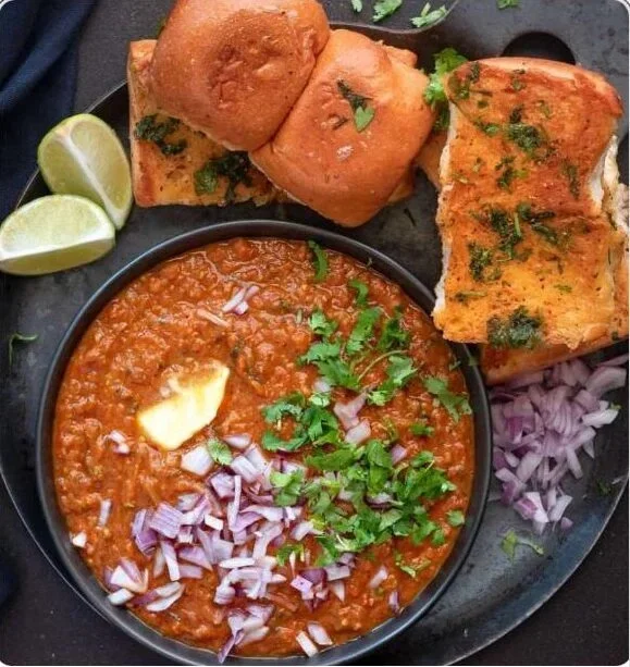 A plate of Indian food featuring a bowl of spicy tomato-based curry with garnished cilantro and onions, a piece of buttered bread, and three fried bread rolls with chopped herbs, served with lime wedges.