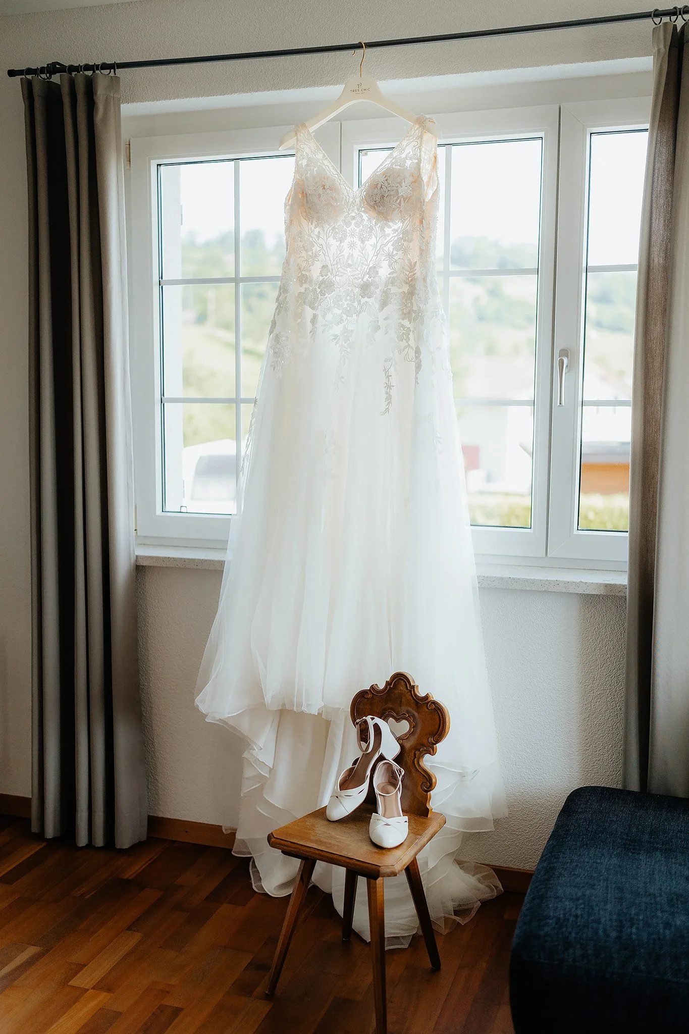 Ein weißes Brautkleid mit Blumenstickerei hängt vor einem Fenster, darunter ein Paar weiße Hochzeitsschuhe auf einem kleinen Holzstuhl, daneben ein Holzstuhl mit einer Herzgravur