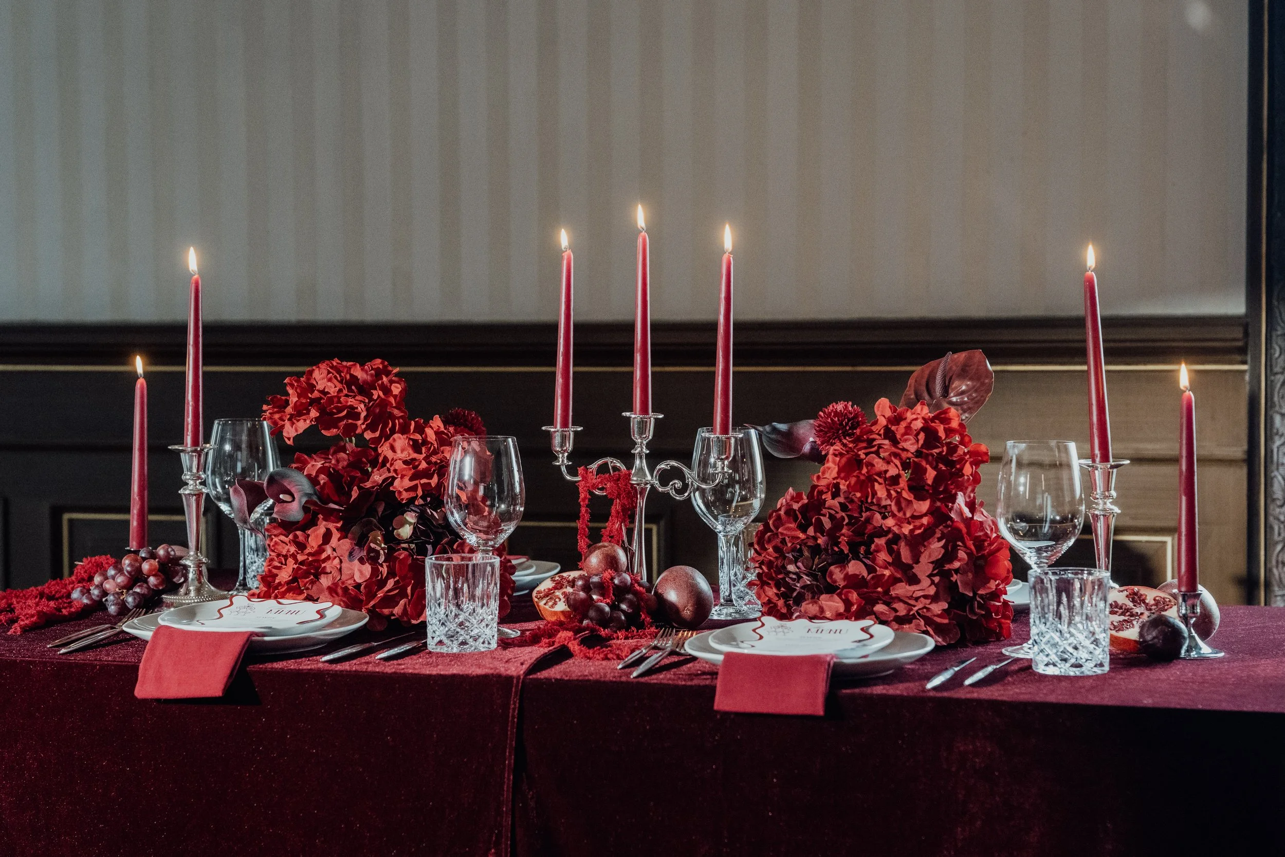 Ein festlich gedeckter Tisch mit roten Blumen, Kerzen, Gläsern und Tellern für eine Feier im eleganten Raum.