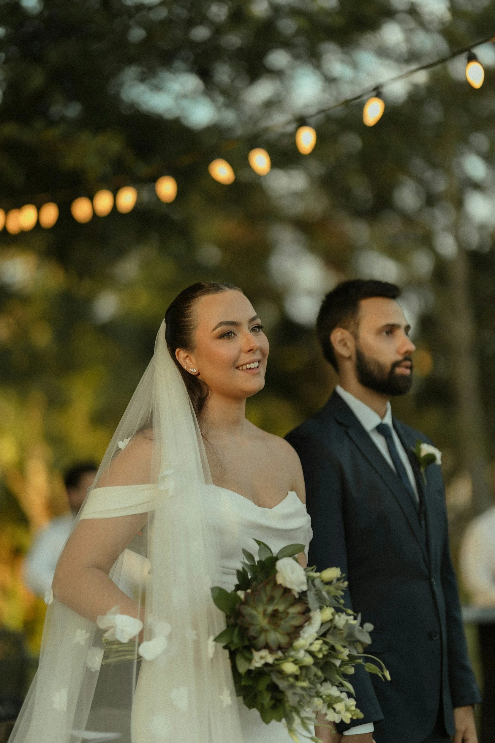 Ein Brautpaar bei einer Hochzeit im Freien, bei Sonnenuntergang, mit Lichterketten im Hintergrund.
