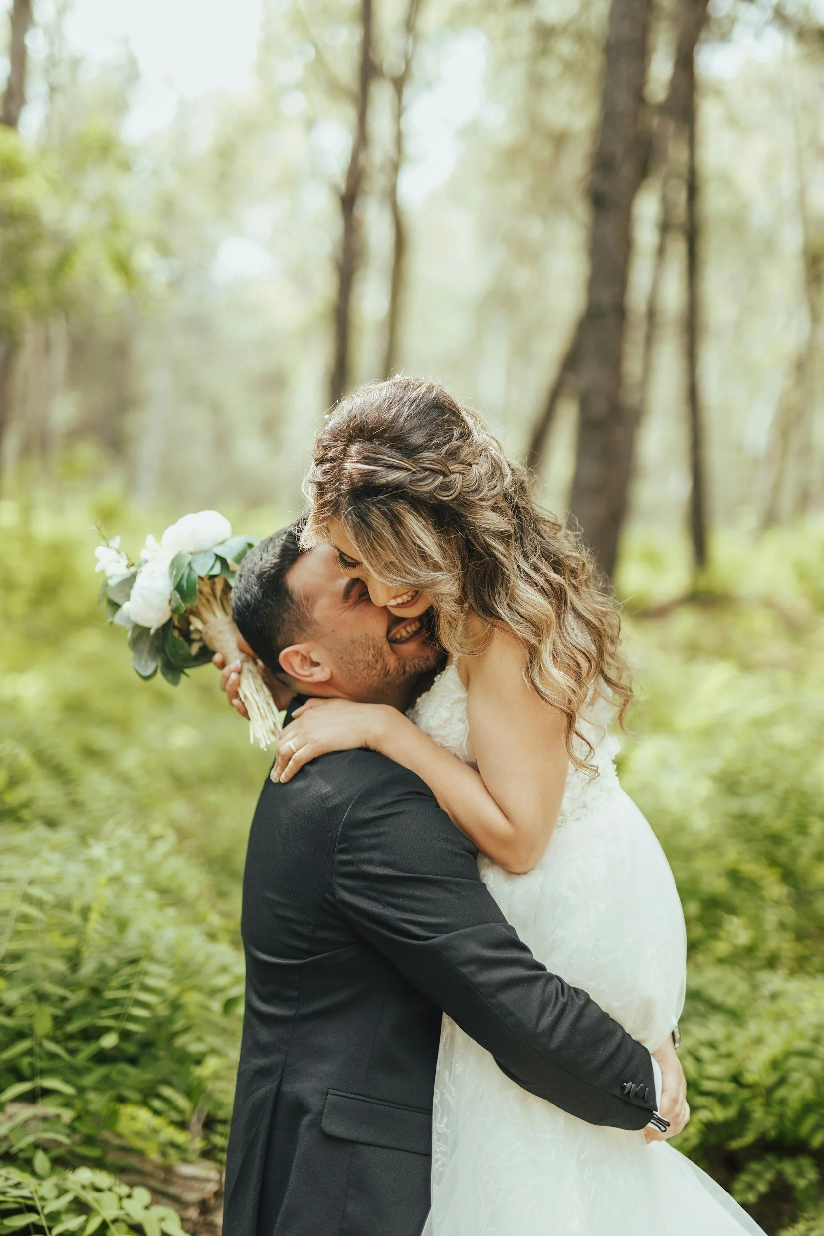 Ein glückliches Brautpaar umarmt sich in einem Wald. Der Bräutigam trägt einen schwarzen Anzug, die Braut ein weißes Hochzeitskleid. Sie lachen und sind sehr verliebt.