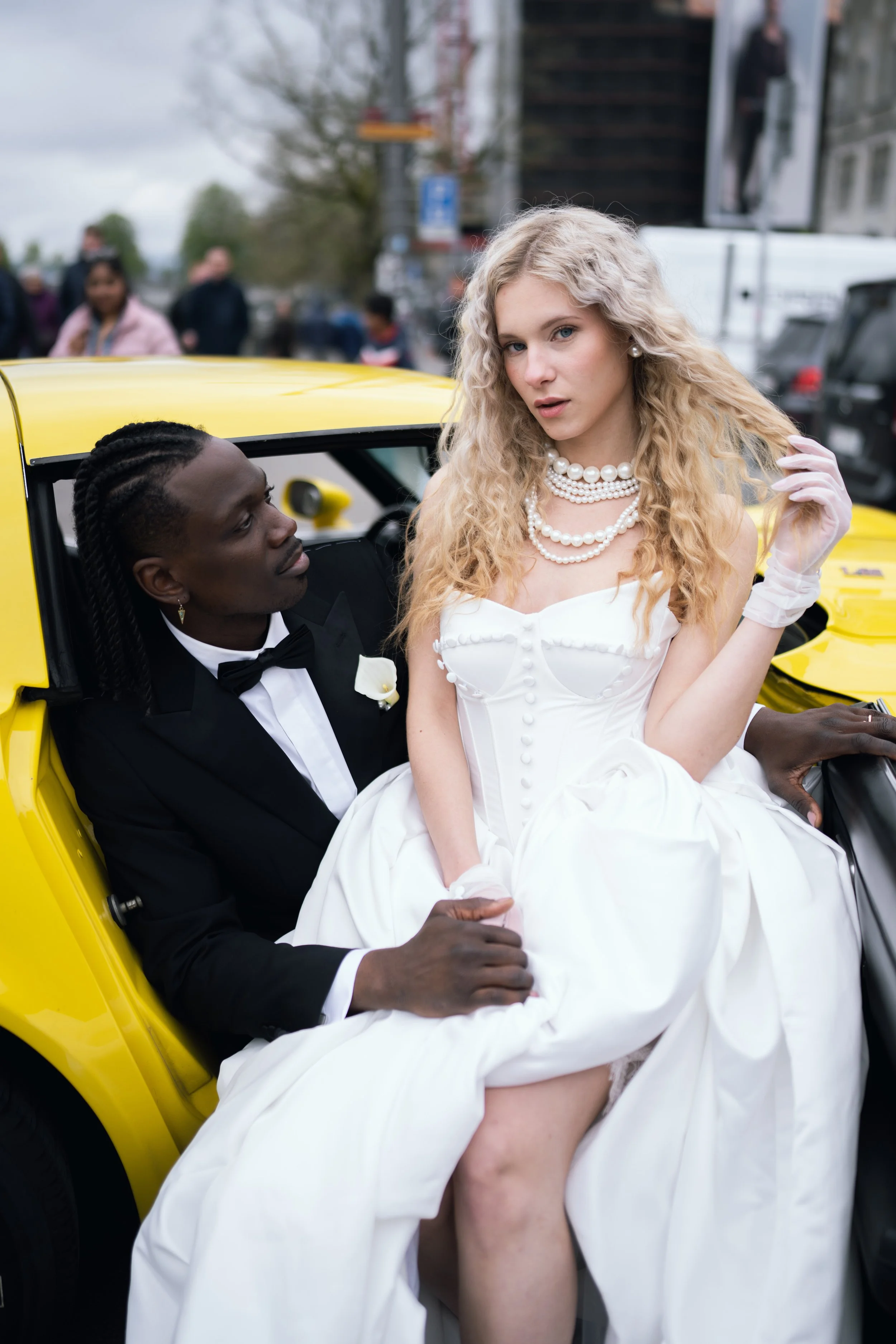 Ein Mann in schwarzem Anzug und Tuxedo sitzt in einem gelben Auto, während eine Frau in einem weißen Hochzeitskleid ihn auf dem Schoß hat. Sie scheinen in einer Stadt auf der Straße zu sein, während im Hintergrund andere Menschen vorbeigehen.