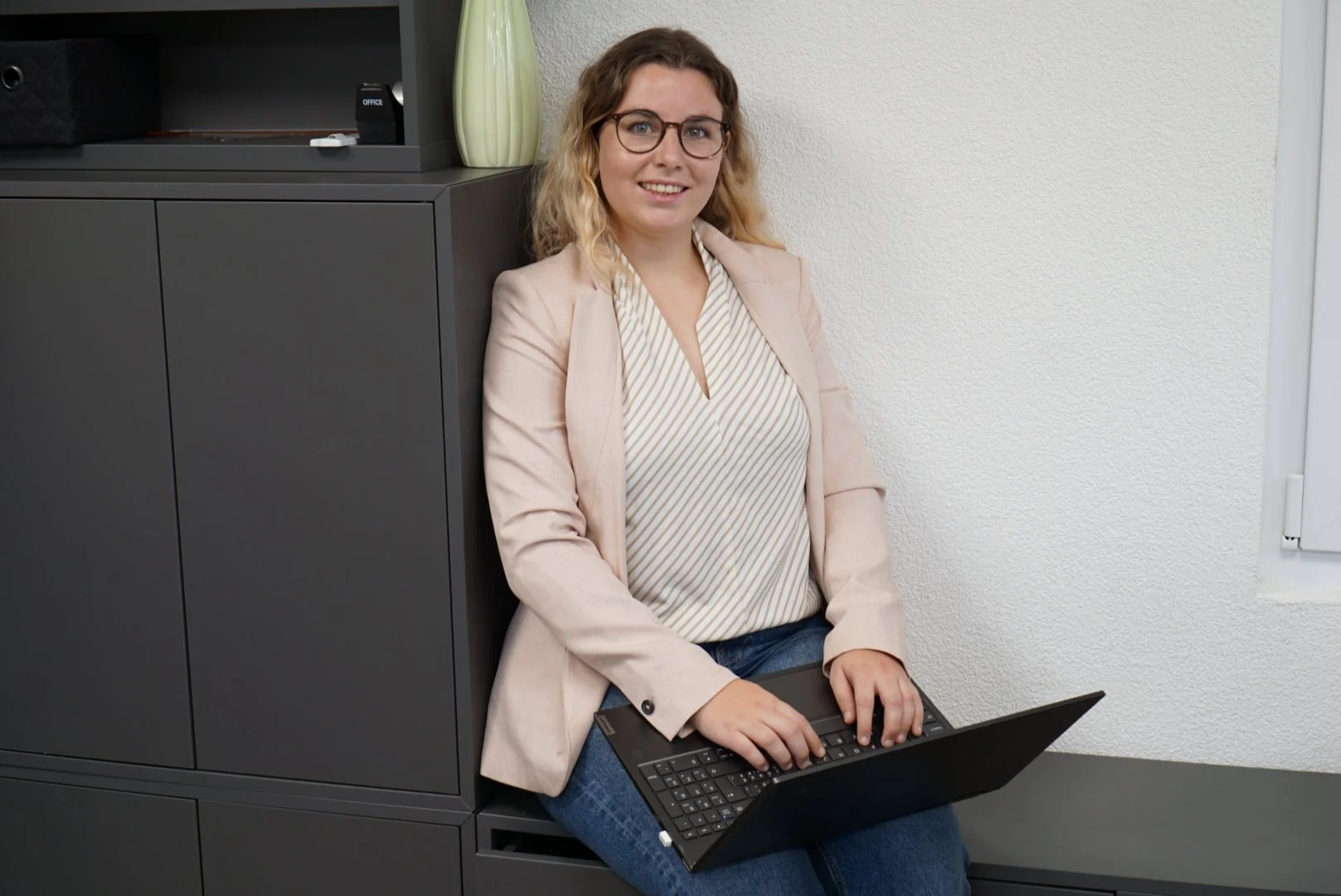 Frau mit Brille sitzt auf einem schwarzen Stuhl in einem Büro und arbeitet an einem Laptop.