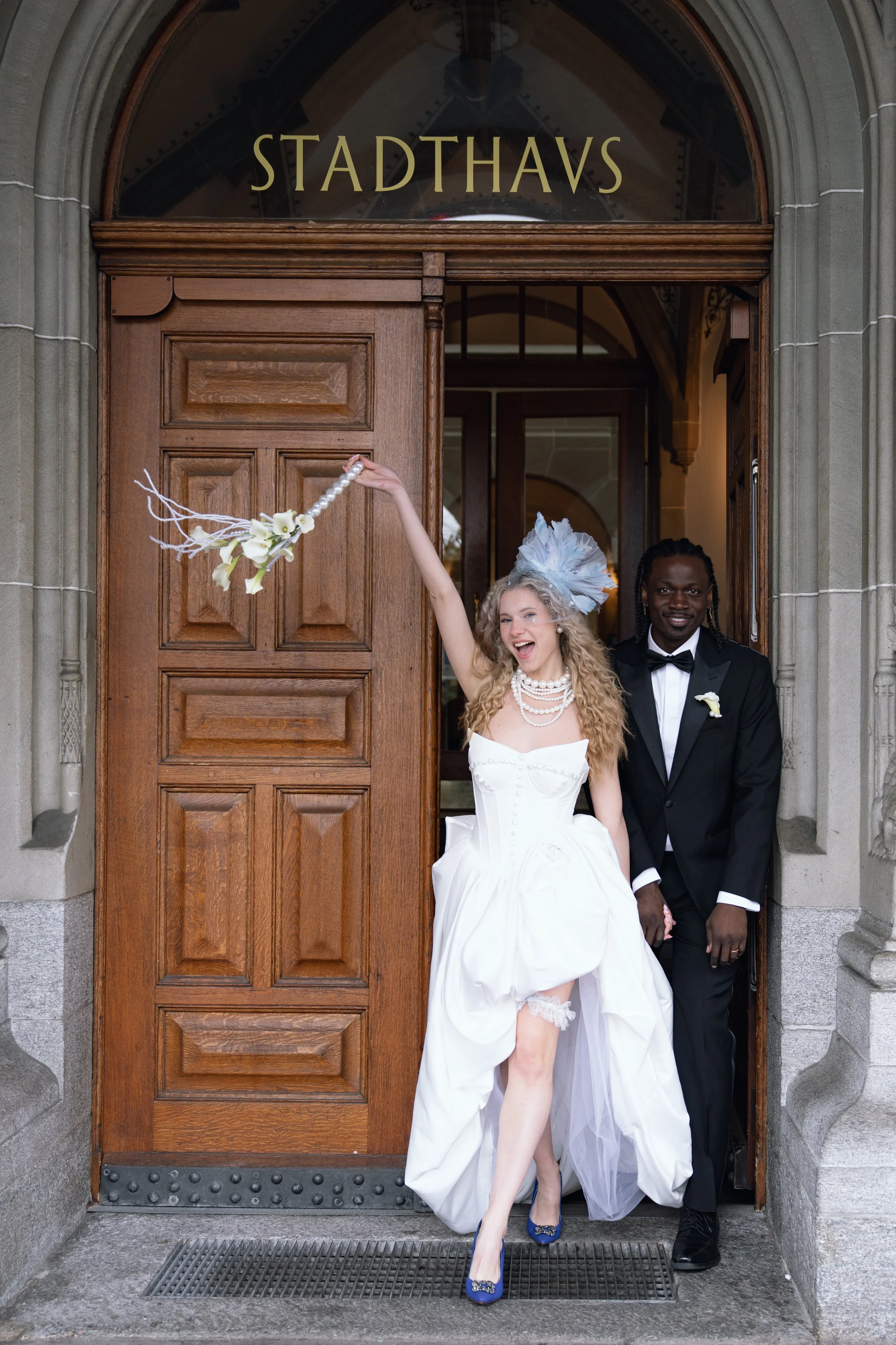 Ein Paar in Hochzeitskleidung steht vor einem Holztürbogen mit der Aufschrift 'Stadthavs'. Die Braut trägt ein weißes Hochzeitskleid, einen Perlen-Schmuck und blaue High Heels, während der Bräutigam einen schwarzen Anzug mit Fliege trägt. Beide sind 