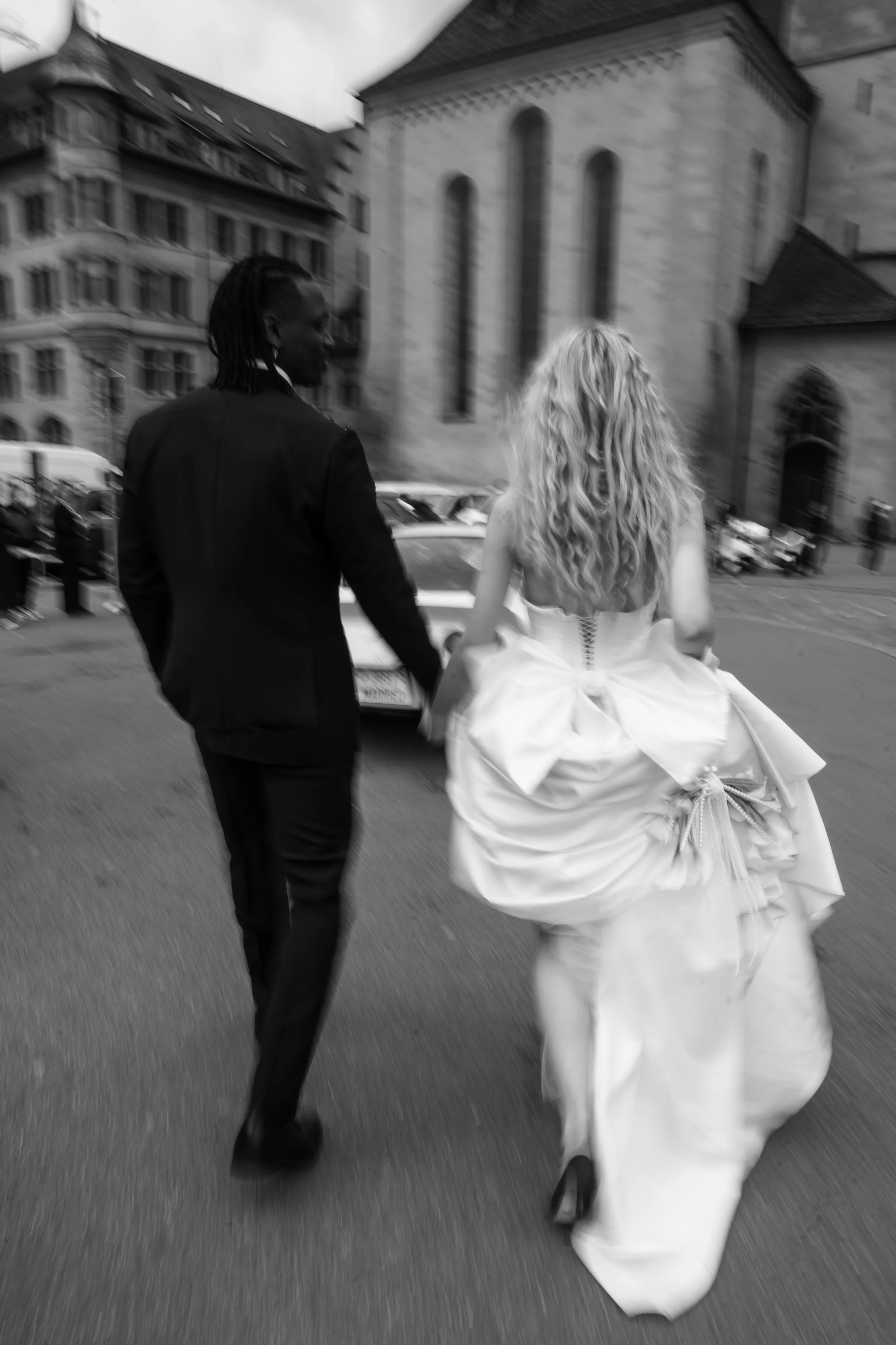 Schwarz-Weiß-Foto eines Paares in formeller Kleidung, das Hand in Hand durch eine Stadt läuft, mit einem alten Gebäude im Hintergrund.