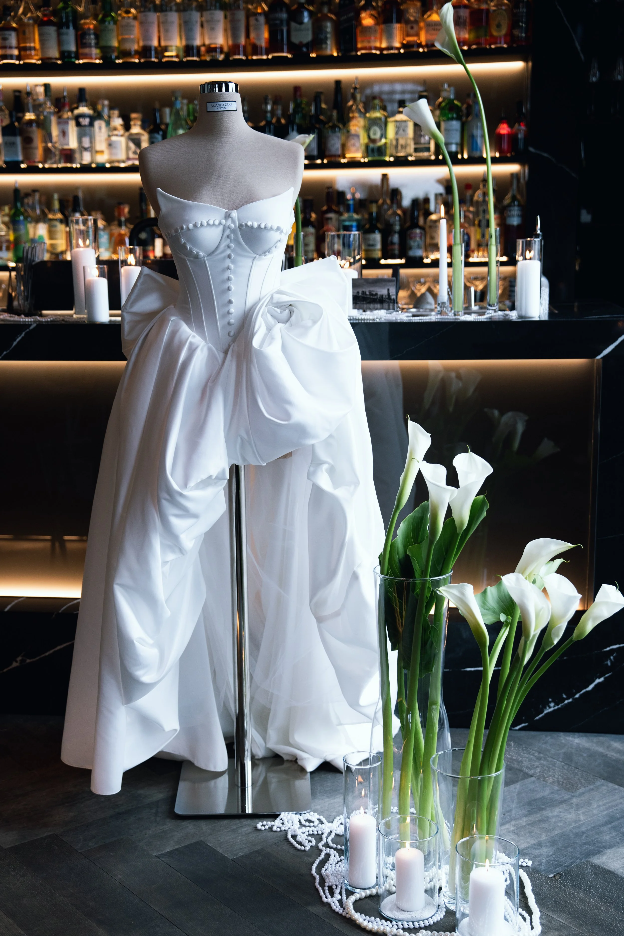 Hochzeitsdekoration mit einem weißen Brautkleid auf einem Mannequin, umgeben von weißen Blumen, Kerzen und Dekorationsmaterial in einem stilvollen Raum mit Bar im Hintergrund.