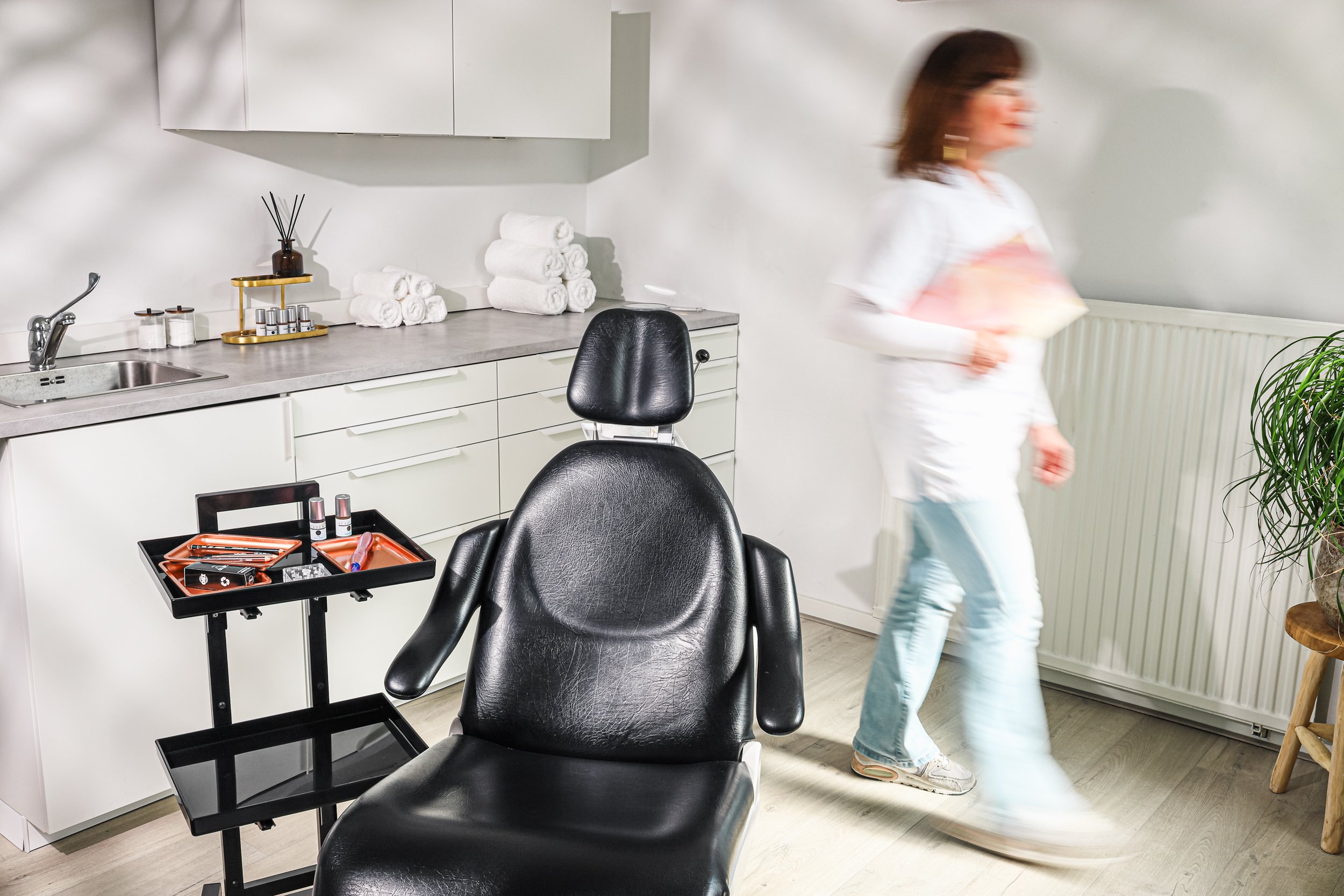 A woman walking past a makeup or beauty treatment room with a black chair, a cart with makeup supplies, and neatly stacked white towels on a countertop.