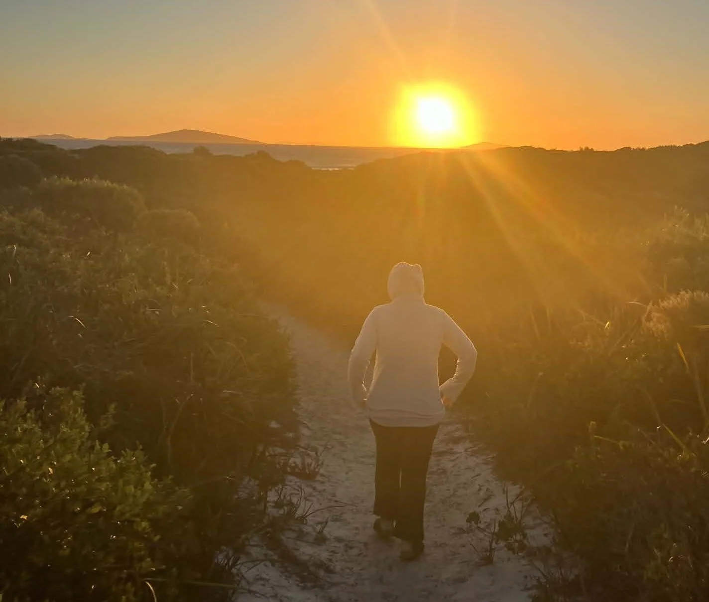 A person wearing a white hoodie and dark pants walking along a path through a lush green landscape at sunset, with the sun low on the horizon casting golden light and rays across the sky.