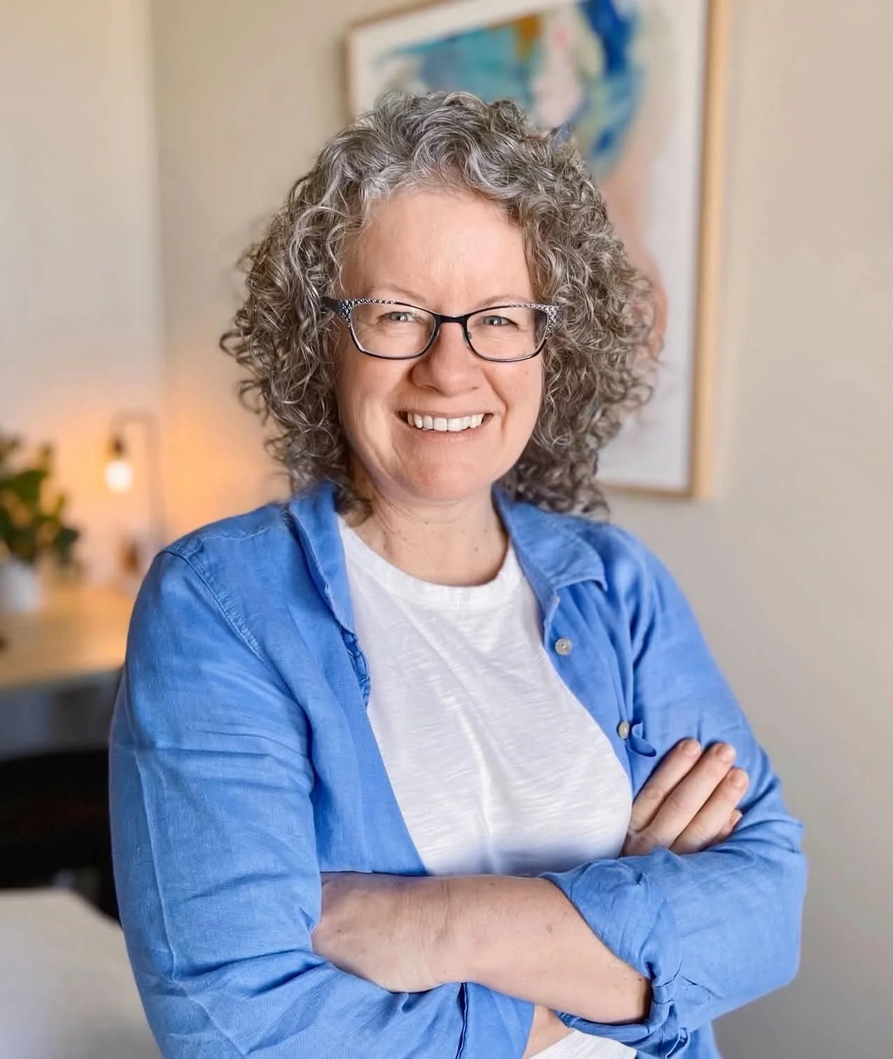 A woman with curly gray hair and glasses smiling, wearing a blue shirt over a white t-shirt, standing with arms crossed in a cozy, well-lit room decorated with a colorful abstract painting and a small plant in the background.
