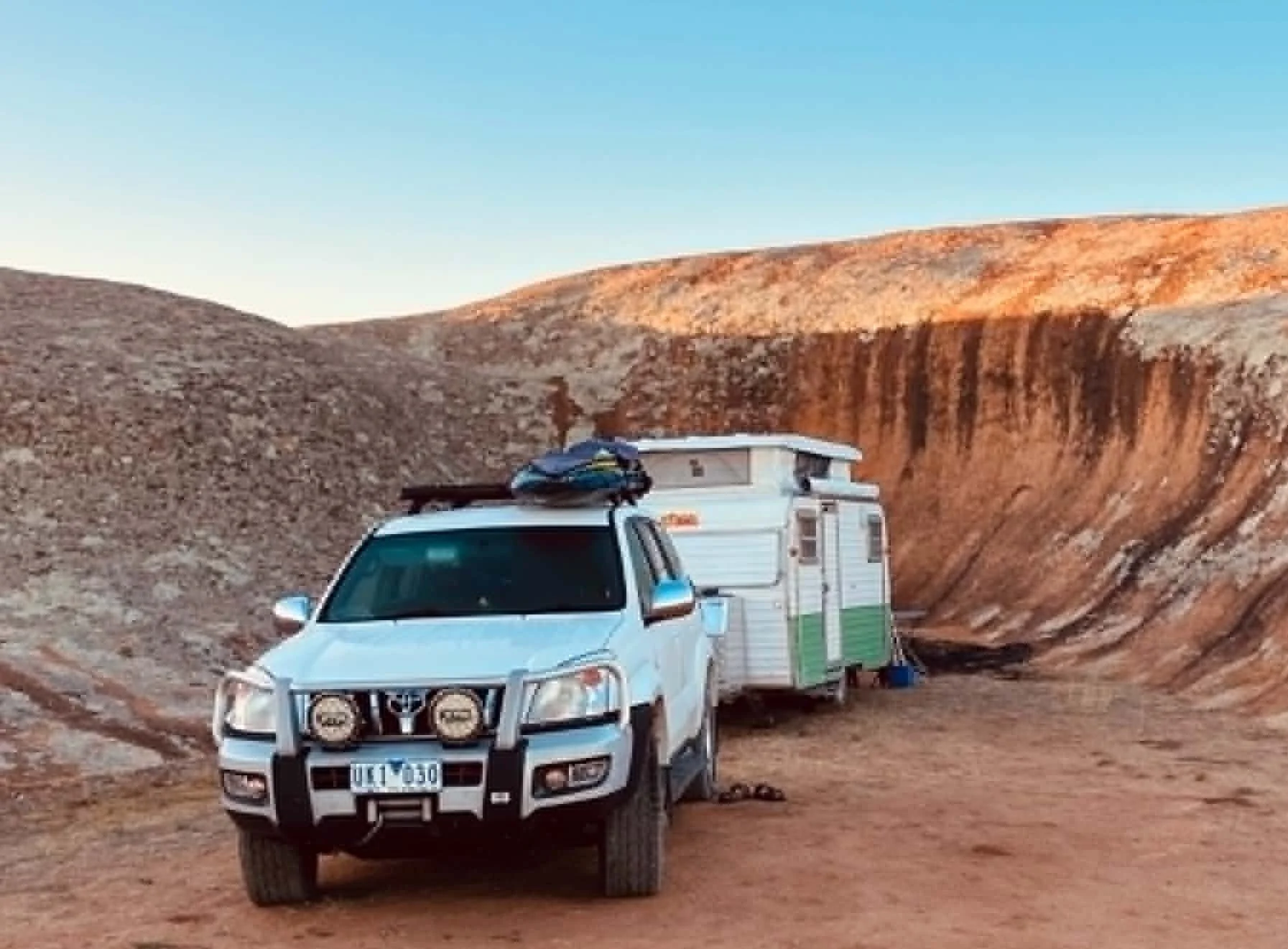 A white pickup truck with a black front bumper and four round off-road lights parked in a desert canyon. Behind it is a small white camper trailer with green accents. The canyon walls are reddish-brown and the sky is clear blue.