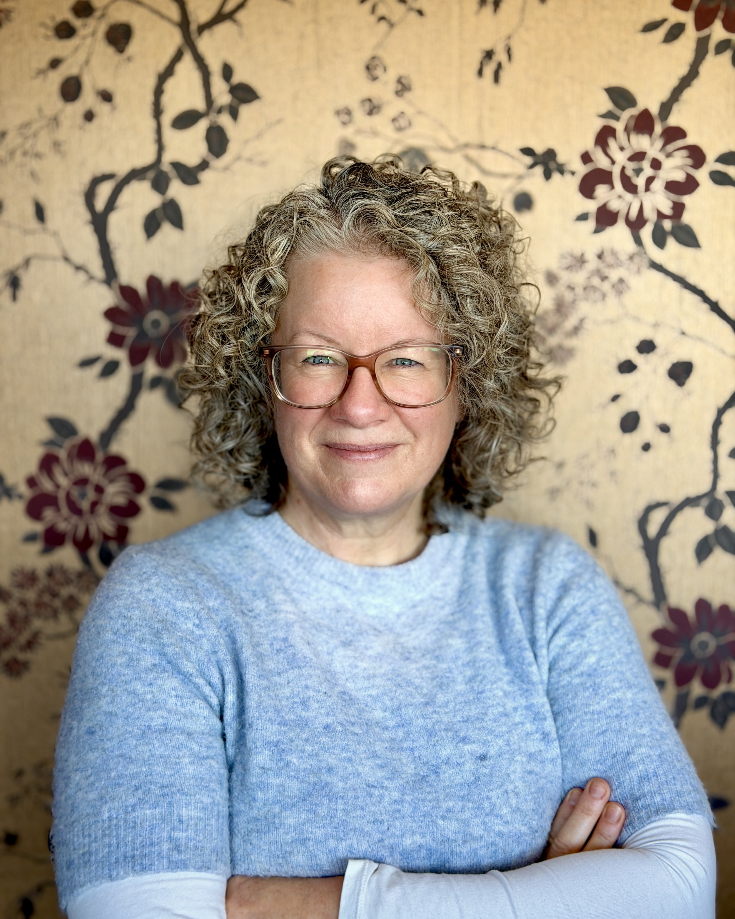 A woman with curly gray hair wearing glasses and a blue sweater, standing against a floral patterned background with large red, black, and cream flowers.